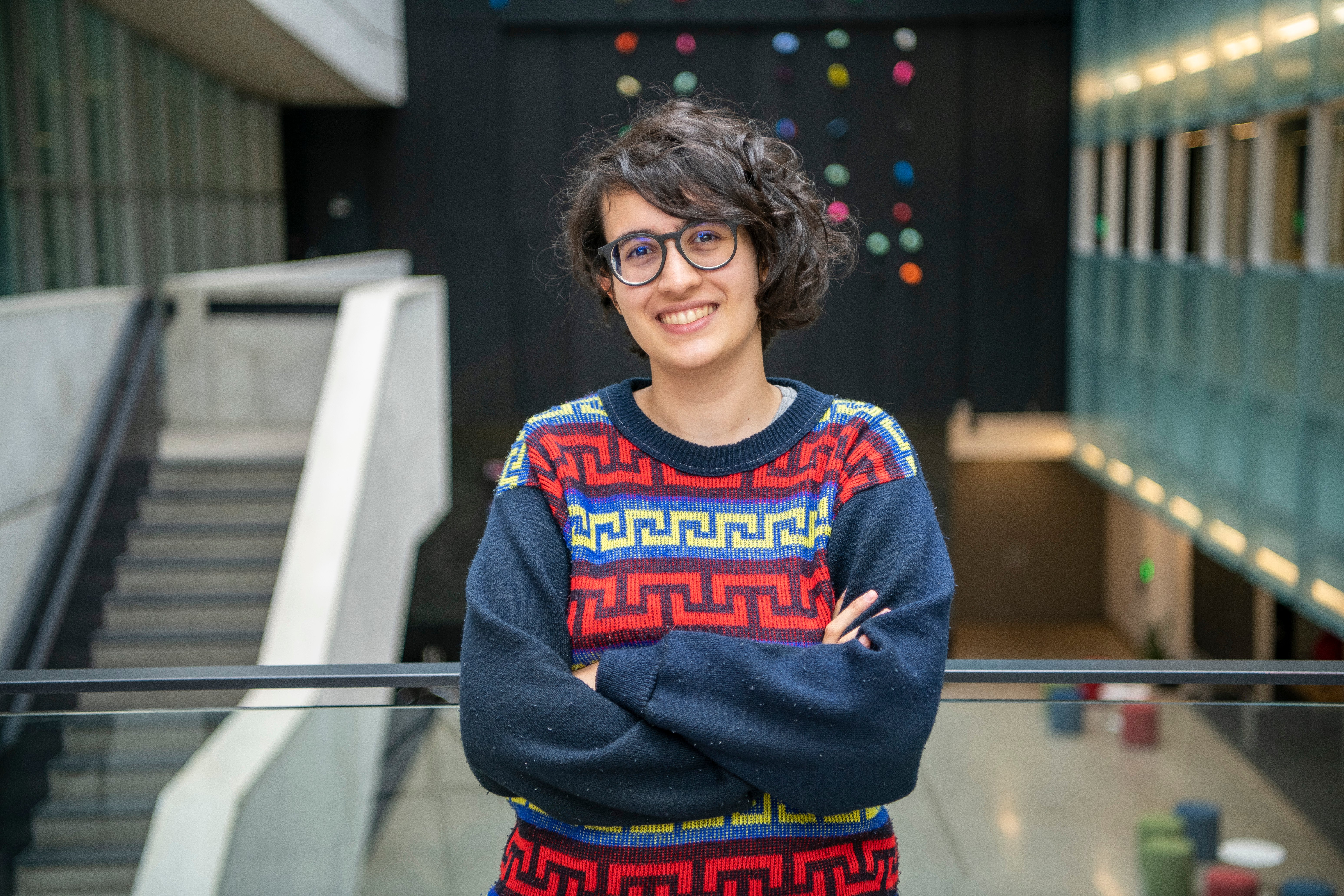 Woman with short dark curly hair and glasses wearing a blue pattern sweater standing in an atrium