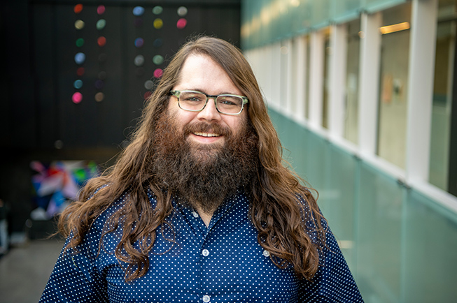 Portrait of a man with long hair and a beard wearing glasses and a blue shirt standing in an atrium