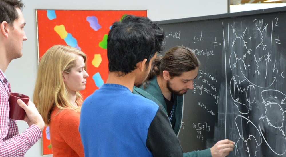 Three men and a woman standing at a chalkboard working on equations