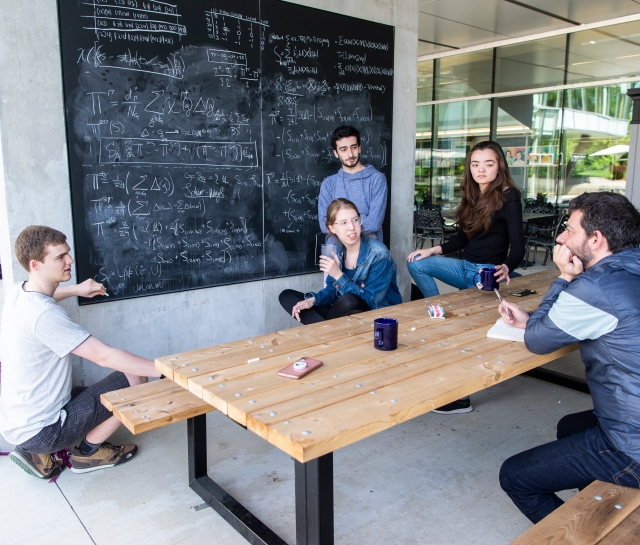 Group of students sitting at a picnic table in front of a blackboard discussing science