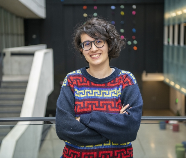 Woman with short dark curly hair and glasses wearing a blue pattern sweater standing in an atrium