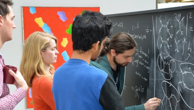 Three men and a woman standing at a chalkboard working on equations