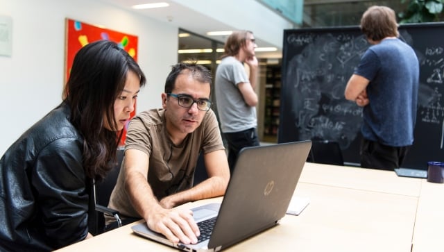 Man and woman working at a computer with two men working at a blackboard in the background