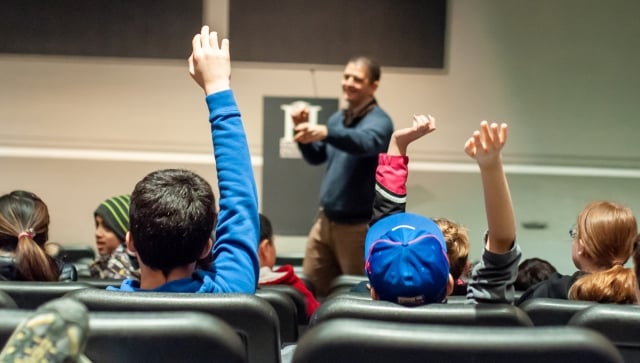 Kids in a lecture hall, raising their hands, asking questions