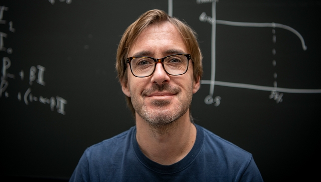 Portrait of a man wearing dark glasses and a blue shirt standing in front of a chalkboard of equations