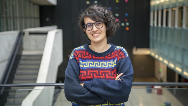 Woman with short dark curly hair and glasses wearing a blue pattern sweater standing in an atrium