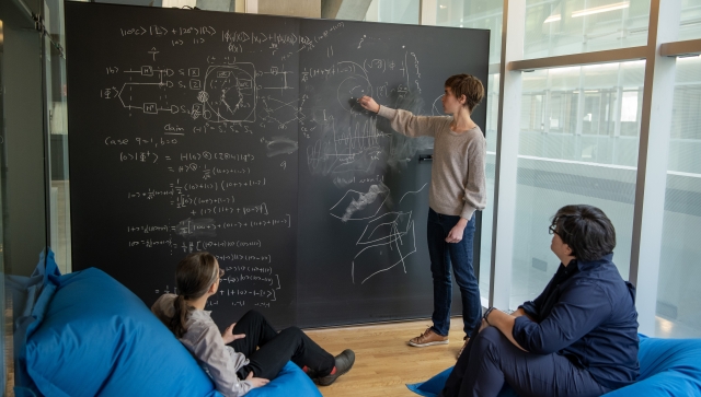 Group of women around a chalkboard, one standing, two sitting on bean bag chairs, talking physics