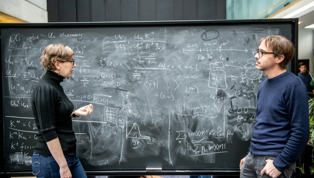 A woman and man standing at a blackboard together discussing physics and writing equations
