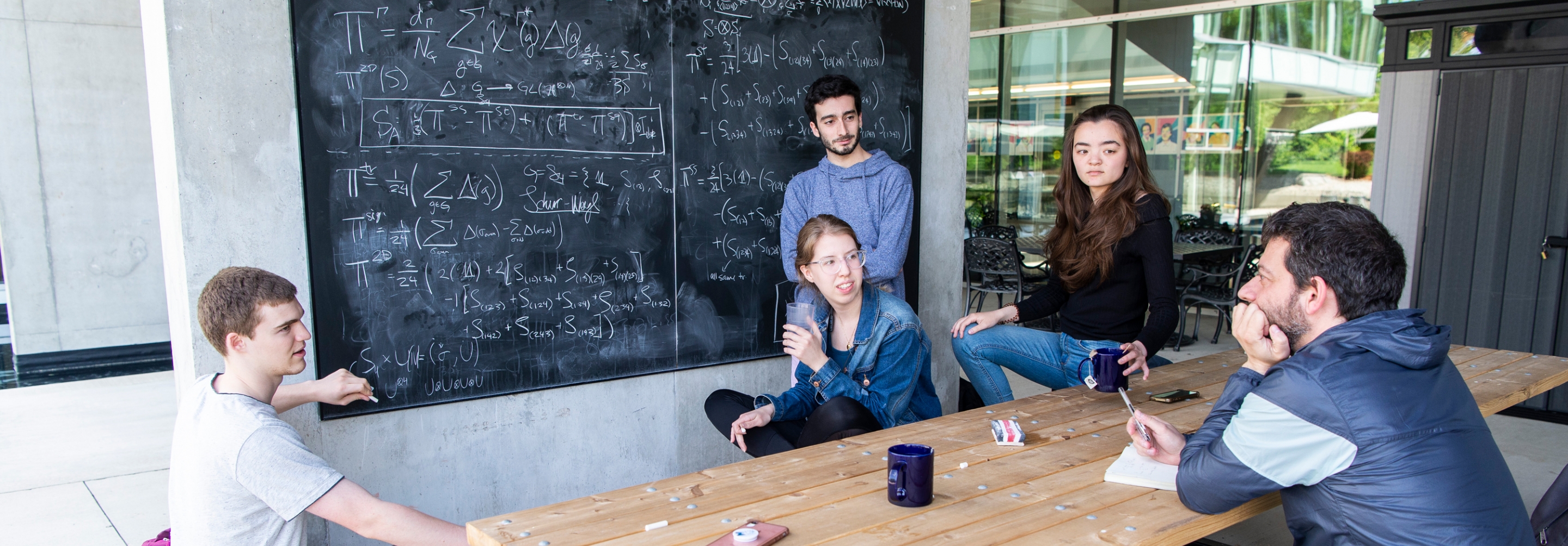 Group of students sitting at a picnic table in front of a blackboard discussing science