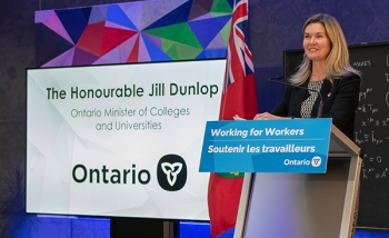Jill Dunlop stands at a podium with a "Working for Workers" sign making a speech beside a banner saying she is Ontario Minister of Colleges and Universities