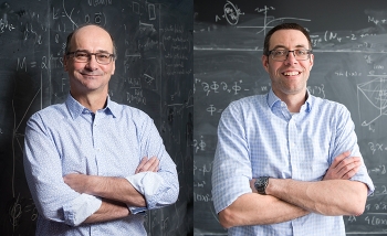 Two men wearing light blue shirts standing in front of blackboard of equations