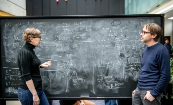 A woman and man standing at a blackboard together discussing physics and writing equations