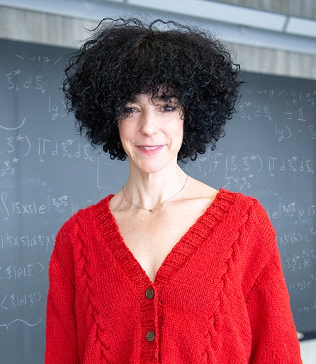 Portrait of a woman with dark hair and a red cardigan standing in front of a blackboard with equations