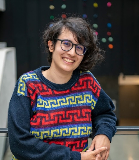 A young woman with short curly dark hair and glasses with a patterned sweater standing in an atrium